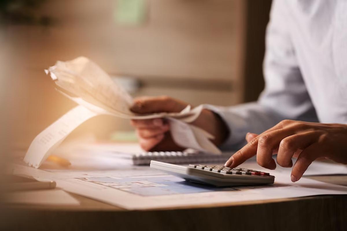 Foto mostra homem fazendo contas em uma mesa, com uma calculadora e folhas em mão. Imagem ilustrativa da situação financeira dos signos durante o mês de agosto.
