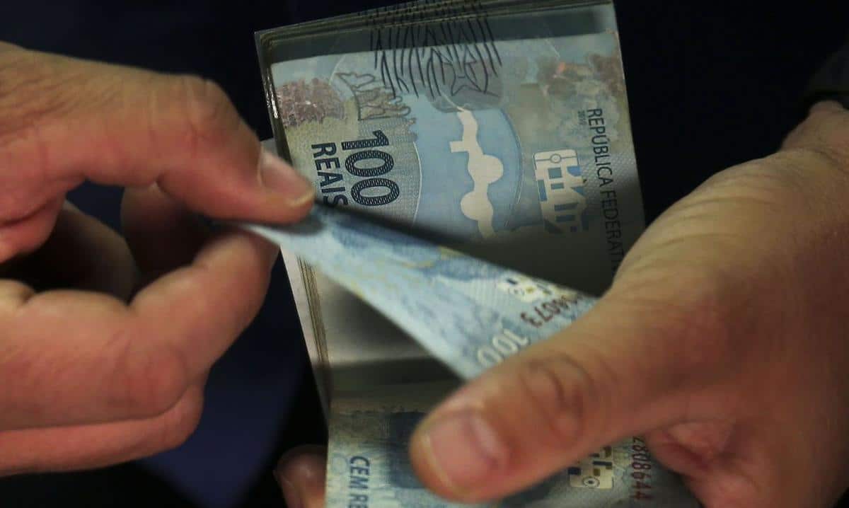 Foto mostra duas mãos folheando notas de R$ 100. Imagem ilustrativa do dinheiro que pode chegar para pessoas de certos signos.