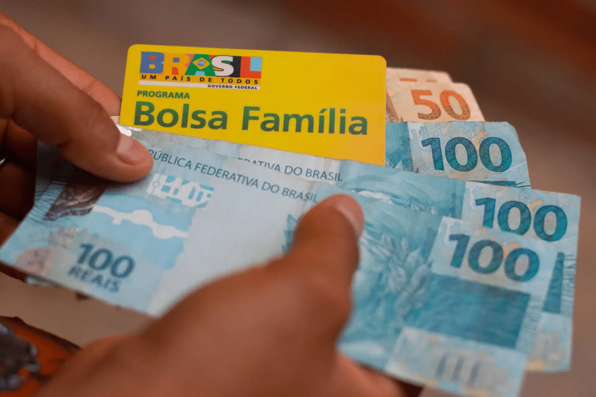 Foto mostra pessoa segurando cartão do Bolsa Família e diversas notas de R$ 50 e R$ 100.